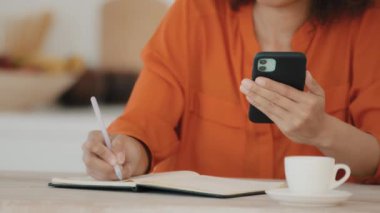 Unrecognizable African woman in orange blouse sits at table at home in office in cafe with cup of coffee holds mobile phone writes in notebook notes writing information online learning business deal