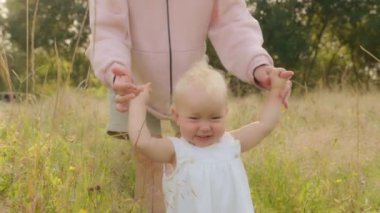 Happy toddler smiling walks through meadow in green high grass holding unrecognizable mother by hand. Mom holding kid daughter baby helps teaches walking in park. First steps of little baby outdoors
