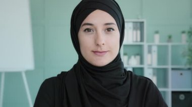 Female portrait happy Arabian 20s girl in black hijab Islamic Muslim woman posing indoors looking at camera looking at camera carefree friendly face businesswoman smiling lady leader boss headshot