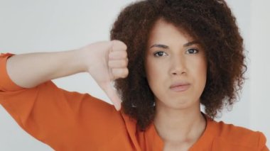 Headshot portrait indoor displeased millennial woman with curly hair looking at camera showing thumbs down rejection finger dislike. Dissatisfied girl disapproval disappointment face reaction bad idea