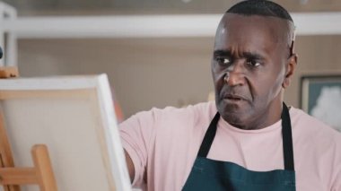 Konsantre olmuş, ciddi, dalgın, yaşlı, Afrikalı sanatçı, yaşlı adam ressam, boya fırçasıyla yağlı akrilik resim çiziyor, sanat fikrini düşünerek tuvalde şaheser yaratıyor.