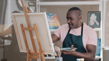 Talented elderly African American male artist old 50s biracial painter in apron works on abstract oil acrylic painting busy concentrated with paint brush creates modern masterpiece picture on canvas