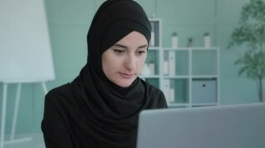 Busy serious Islamic Muslim businesswoman typing laptop unexpected call answer phone calling talking smartphone angry discussing problems conflict quarrel say no disagree business trouble at work