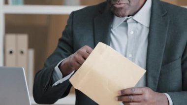 Amazed shocked African old 50s man in office receive mail correspondence senior mature businessman opening paper envelope reading letter with good news offer celebrate victory win approval bonus prize