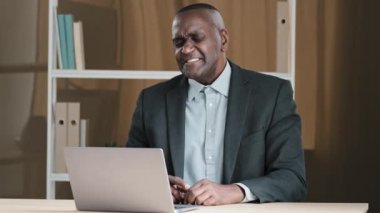 African American businessman mature man boss leader manager banker old 60s worker sits in office table listen music with laptop singing dancing enjoy online victory winning celebrate dance to song
