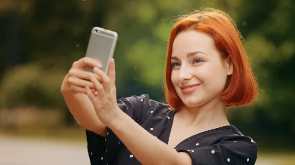 Portrait stylish beautiful girl caucasian attractive redhead woman blogger female model stands outside in park holds smartphone takes selfie photo on mobile camera records vlog filming video for blog