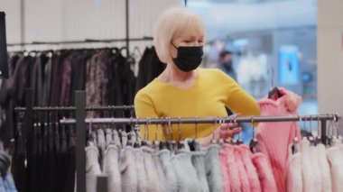 Mature woman customer in protective mask chooses colorful trendy sweater in clothing store elderly female shopper looking for new outfit in modern boutique in search of stylish clothes renews wardrobe