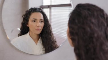 Close-up serious pensive young hispanic woman looking in mirror in bathroom contemplating future cute curly brunette girl in bathrobe admires reflection natural beauty daily morning routine concept