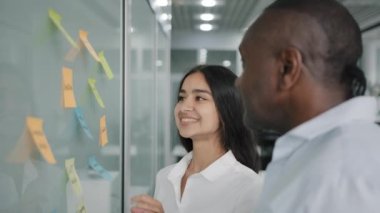 Business team colleagues diverse co-workers discusses the project together choose sticky notes paper memo sticks on a wall board african man plans work process with arabic woman brainstorming session