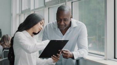 Diverse african and arabian colleagues multiracial people business partners stand near window discussing paper graphics strategic plans reading documents in tablet folder for paperwork documentation