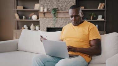 Concentrated adult african businessman american senior man writer freelancer analyst holds papers utility bills documents makes remote payment e-pay using banking app typing text on computer keyboard