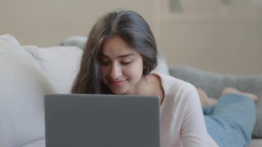 Carefree woman lying on sofa working on laptop searching online information playing computer game. Arabian middle-eastern female freelancer student search job vacancy in internet e-learning education