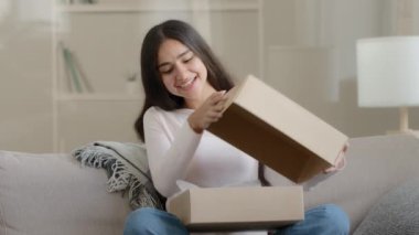 Surprised arab latin woman satisfied customer shopaholic sits on couch at home open delivery parcel postal carton box unpack present gift look at shipping goods purchases from internet clothing store