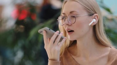 Close up emotional girl use virtual assistant speak in microphone of phone caucasian young lady with glasses talk on speakerphone record audio message using wireless headphones telephone communication