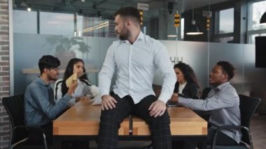 Chaotic co-workers in background arguing multiracial colleagues in boardroom young frustrated man worker pointing head making finger gun irritated tired employer sitting on table pretending headshot
