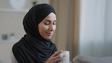 Portrait of happy young muslim woman resting at home drinking hot tea enjoying weekend smiling girl in hijab dreaming about future feels positive emotions lady looks into distance relaxing in solitude