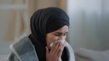 Close up sick upset young girl in hijab sneeze wipe nose with paper napkin sad unhealthy muslim woman drinking hot tea suffering from seasonal cold feel virus flu symptoms coronavirus epidemic concept