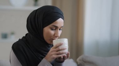 Carefree joyful muslim girl in hijab drinking tea relaxing in room alone look thoughtfully into distance happy islamic young woman enjoy rest weekend dream of future feel stress-free home cozy concept
