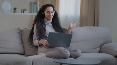 Joyful successful hispanic girl sit on sofa make video call communicate remote in online conference use laptop and headphones happy smiling young lady freelancer working at home concept modern devices