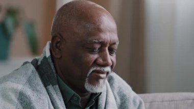 Close up ill sick elderly african american man covered with warm blanket sitting on couch in living room senior retiree holding cup drinking tea at home pensioner isolation in apartment resting alone