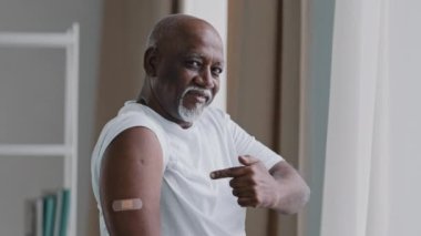 Happy elderly african american man show adhesive bandage on shoulder after vaccination smiling grandpa looking at camera satisfied immunization point finger at medical patch disease protection concept