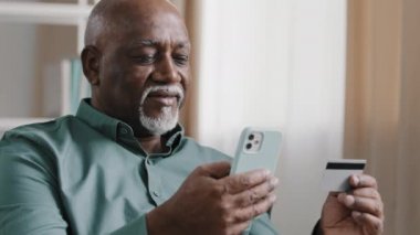 Smiling elderly mature man hold telephone and credit card male customer make online purchase pay bills use bank app enter number for transaction happy african american enjoy modern tech secure payment
