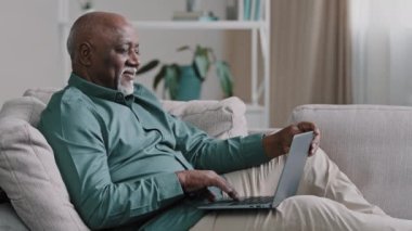 Happy mature pensioner sitting on sofa making internet order using modern laptop african american shopper browsing website smiling carefree elderly grandpa pays bank bills remote communication concept