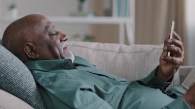 Close up relaxed lying on couch elderly african american man making video call use smartphone mature smiling grandfather holding mobile phone answer talking share good news waving hand show thumbs up