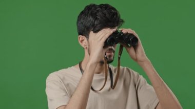 Close up happy hispanic guy using binoculars indian cheerful tourist looking adventure studio green background portrait friendly young arab man intently peep looking at camera journey safari concept