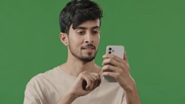 Portrait close-up arabian young man in studio on green background holding phone happy smiling guy browsing social networks make online order read news using modern gadget male customer shopping remote