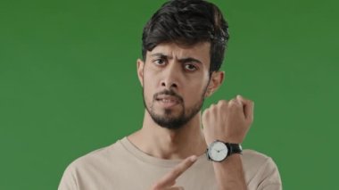 Portrait serious indian young man standing over green background angry nervous male shake head close up dissatisfied frustrated hispanic guy pointing finger wrist watch late missed wasted time concept