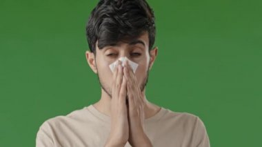Close-up in studio with green background sick hispanic guy sneezes wipes with paper napkin young man suffering from seasonal allergies runny nose arab male feel uncomfortable flu symptom virus illness