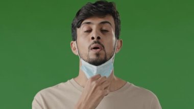 Tired young hispanic man in medical mask on green background in studio arab guy take off respirator enjoy fresh air smiling male inhale deeply breathing freedom exhale with relief break lack of oxygen