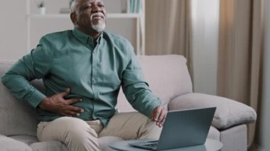 Older african american male freelancer mature pensioner sitting at home office on couch using laptop feel acute stomach pain discomfort indigestion digestive problems sharp ache in abdomen bad feeling
