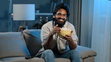 Bearded man in glasses indian male bachelor after work sits on sofa in dark living room turns on TV with remote control choosing channel watching online movie film program eating popcorn. High quality