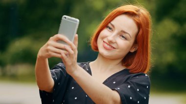Kafkasyalı güzel, kızıl saçlı, stil sahibi kadın blog yazarı selfie çekiyor, kendine hayranlık duyuyor modern akıllı telefon kamerasından video yaşam tarzı blogu parkta çevrimiçi çeviri sağlıyor.
