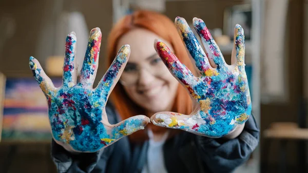 Close-up happy funy woman with red hair painter girl artist in glasses looks at camera stretches forward hands showing dirty palms smeared in acrylic paint laughs inspired by drawing decor design art