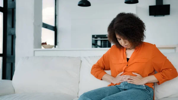 Stressed African American woman sitting on sofa touching belly ill sick biracial curly girl suffering from stomachache. Unhappy lady feeling ache menstrual painful unwell upset with abdominal pain