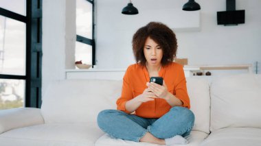 Coşkulu Afrikalı Amerikalı kadın akıllı telefon yazma fırsatı buldu. Mobil uygulamada para ödülü kazandı. Evdeki koltukta oturdu. Kazanan mutlu kadın zaferi kutluyor.