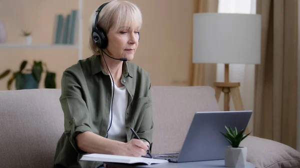 Senior businesswoman in headphones sitting on couch listen online training webinar distant e-learning 60s woman business class video chat call conference on laptop computer remote working writing note