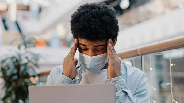 African american sad business man guy student wearing medical mask working with laptop in corporate office holding head feeling headache suffering pressure migraine coronavirus symptoms overwork. High