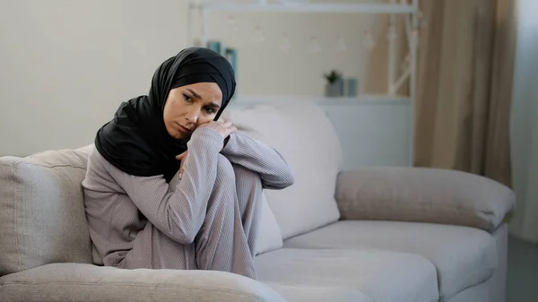 Anxious sad young woman in black hijab sitting on couch in living room suffering from psychological problem trouble at home frustrated muslim girl feeling stress depressed lost in negative thoughts