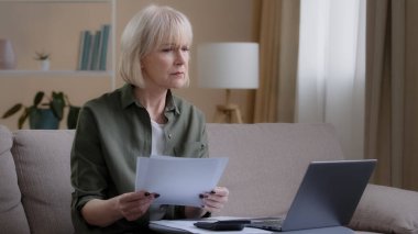 Busy 50s homeowner housewife makes woman budget checks monthly income by checking paper notes using app laptop. Middle-aged businesswoman sitting on couch with document working remote at home. High