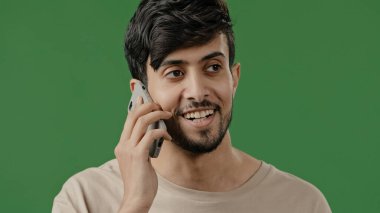 Mutlu gülümseyen genç Arap stüdyoda yeşil arka planda konuşan akıllı telefon kullanan, pozitif adam cep telefonu modern teknoloji kavramında arkadaşlarıyla hoş sohbetler yapıyor.