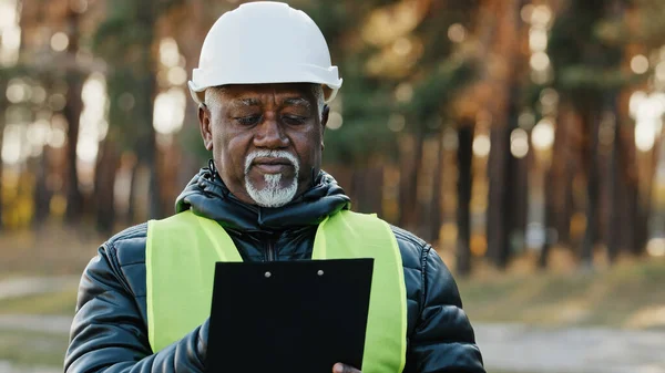 Mature concentrated forestry engineer technician forester in protective helmet stands in park assesses situation makes plan for cutting trees observes reserve writes data takes reforestation measures