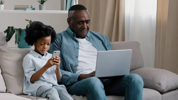 Upset male freelancer sitting on sofa in room with laptop dissatisfied with result loses online game gets bad news little cute daughter sitting next to dad smiling holding mobile phone watching father