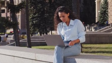 Gülümseyen neşeli, tasasız genç kız öğrenci başarılı iş kadını laptopuyla şehirde oturmuş sohbet ediyor komik videolar seyrederek iyi haberler alıyor.