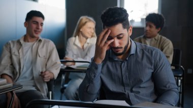 Sad frustrated arab student sitting in class at desk alone suffering from mistreatment by classmates racial discrimination feels victim of bullying violent students bully ridicule gossip behind back