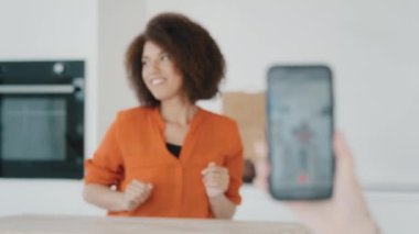 Blurred view African woman dancing in kitchen. Close up female hand holding mobile phone taking video making photo online smartphone screen with curly girl dancing funny moving having fun at home