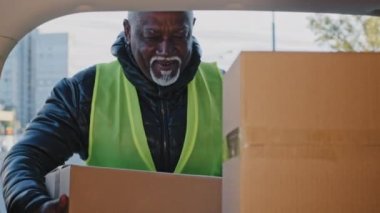 Close-up happy mature african american man in workwear uniform stacking cardboard boxes courier loader handyman delivery worker doing quality job loading goods into truck looking at camera smiling